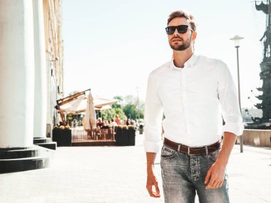 Yakışıklı, gülümseyen stil sahibi Lambersex modelinin portresi. Beyaz gömlek giymiş modern bir adam. Sokakta güneş gözlükleriyle poz veren modacı erkek. Gün batımında dışarıda. 