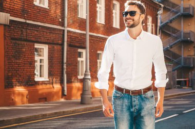Yakışıklı, gülümseyen stil sahibi Lambersex modelinin portresi. Beyaz gömlek giymiş modern bir adam. Sokakta güneş gözlükleriyle poz veren modacı erkek. Gün batımında dışarıda. 
