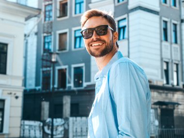 Yakışıklı, güler yüzlü, stil sahibi, lamberseksüel bir modelin portresi. Mavi gömlekli modern bir adam. Moda erkeği güneş gözlükleriyle gökdelenin yanında poz veriyor. Gün batımında dışarıda. 