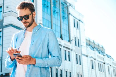 Yakışıklı, güler yüzlü, stil sahibi, lamberseksüel model. Mavi gömlekli, modern bir adam. Sokakta güneş gözlükleriyle poz veren modacı erkek. Gün batımında dışarıda. Akıllı telefon uygulamaları kullanıyor. SMS yazıyor.