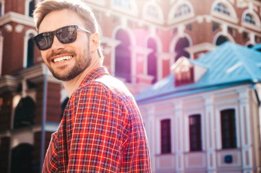 Yakışıklı, şık, havalı, havalı, lamberseksüel model. Kırmızı kareli gömlek giymiş modern bir adam. Moda erkeği güneş gözlükleriyle gökdelenin yanında poz veriyor. Gün batımında dışarıda. 