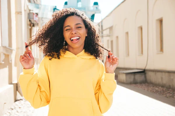 Beautiful black woman with afro curls hairstyle.Smiling hipster model in yellow hoodie. Sexy carefree female posing on the street background. Cheerful and happy