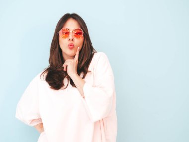 Yeni moda hippi kıyafetleri içinde gülümseyen güzel bir kadın. Stüdyoda açık mavi duvarın yanında poz veren kaygısız seksi kadın. Pozitif model içeride eğleniyor. Neşeli ve mutlu güneş gözlükleriyle