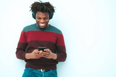 Yakışıklı, güler yüzlü, lamberseksüel model. Şık giyinmiş, traşsız Afrikalı bir erkek. Stüdyoda duvarın yanında saç stili saçı olan, kablosuz kulaklıkla müzik dinleyen moda bir erkek.