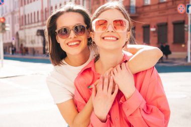 Son moda beyaz tişörtlü ve kot pantolonlu iki güzel, gülümseyen yeniyetme bayan sokak arka planında poz veren kaygısız kadınlar. Pozitif modeller eğleniyor, sarılıyor ve deliriyor.