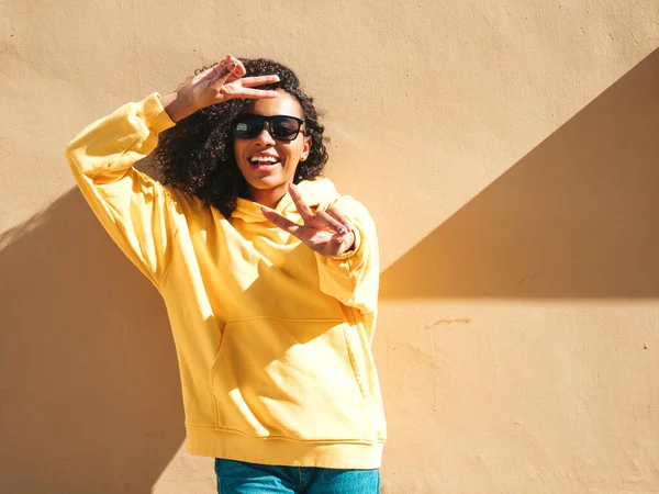 Beautiful black woman with afro curls hairstyle.Smiling hipster model in yellow hoodie. Sexy carefree female posing on the street background near wall in sunglasses.Cheerful and happy.Shows peace sign