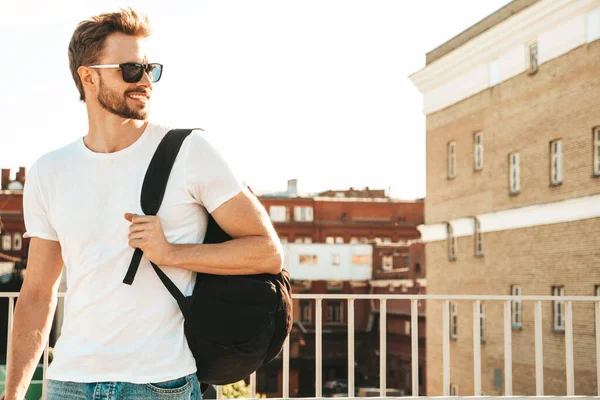 Yakışıklı, güler yüzlü, Lambersex mankeni. Beyaz tişört ve kot giyen şık bir adam. Moda erkek plajda çitlerin yanında güneş gözlüklü okul çantasıyla poz veriyor.