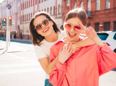 Son moda beyaz tişörtlü ve kot pantolonlu iki güzel, gülümseyen yeniyetme bayan sokak arka planında poz veren kaygısız kadınlar. Pozitif modeller eğleniyor, sarılıyor ve deliriyor.