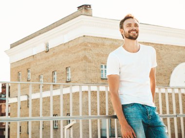 Yakışıklı, gülümseyen hipster Lambersex modelinin portresi. Beyaz tişört ve kot giyen şık bir adam. Moda erkeği, plajdaki çitlerin yanında sokak arkasında poz veriyor. 