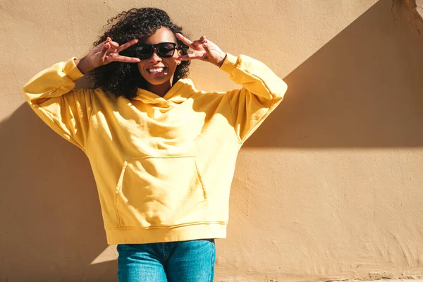 Beautiful black woman with afro curls hairstyle.Smiling hipster model in yellow hoodie. Sexy carefree female posing on the street background in sunglasses. Cheerful and happy.Shows peace sign