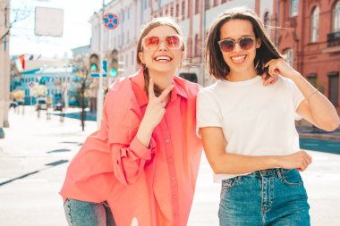 Son moda beyaz tişörtlü ve kot pantolonlu iki güzel, gülümseyen yeniyetme bayan sokak arka planında poz veren kaygısız kadınlar. Pozitif modeller eğleniyor, sarılıyor ve deliriyor.