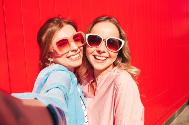 Son moda yaz kıyafetleri içinde iki güzel gülümseyen yeniyetme kadın. Kırmızı renkli duvarın yanında poz veren kaygısız kadınlar. Pozitif modeller eğleniyor, sarılıyor ve Pov 'un selfie' sini çekiyor.
