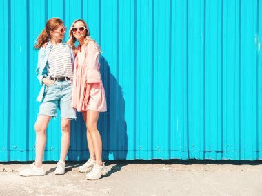 Son moda yaz kıyafetleri ve kotları içinde gülümseyen iki güzel hippi kadın. Mavi duvarın yanında poz veren kaygısız kadınlar. Gün batımında eğlenen, sarılan ve deliren pozitif mankenler.