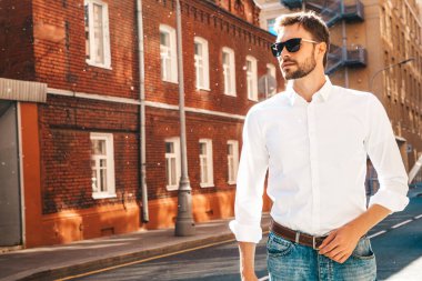 Yakışıklı, kendine güvenen, stil sahibi, Lambersex mankeni portresi. Beyaz gömlek giymiş modern bir adam. Sokakta güneş gözlükleriyle poz veren modacı erkek. Gün batımında dışarıda. 