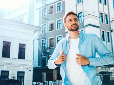 Yakışıklı, güler yüzlü, stil sahibi, lamberseksüel bir modelin portresi. Mavi gömlekli modern bir adam. Moda erkeği, arka planda gökdelenin yanında poz veriyor. Gün batımında dışarıda. 