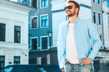 Yakışıklı, güler yüzlü, stil sahibi, lamberseksüel bir modelin portresi. Mavi gömlekli modern bir adam. Moda erkeği güneş gözlükleriyle gökdelenin yanında poz veriyor. Gün batımında dışarıda. 