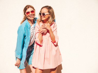 Son moda yaz kıyafetleri ve elbiseleri içinde iki güzel gülümseyen hippi kadın. Beyaz duvarın yanında sokakta poz veren kaygısız kadınlar. Gün batımında eğlenen, sarılan ve deliren pozitif modeller.