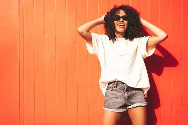 Beautiful black woman with afro curls hairstyle.Smiling hipster model in white t-shirt. Sexy carefree female posing in the street near red wall in sunglasses. Cheerful and happy