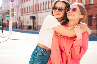 Son moda beyaz tişörtlü ve kot pantolonlu iki güzel, gülümseyen yeniyetme bayan sokak arka planında poz veren kaygısız kadınlar. Pozitif modeller eğleniyor, sarılıyor ve deliriyor.