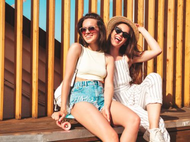 Son moda yaz kıyafetleri içinde iki güzel gülümseyen yeniyetme kadın. Sokakta şapkalı kayla poz veren kaygısız kadınlar. Pozitif saf modeller gün batımında eğleniyorlar, sarılıyorlar ve deliriyorlar.