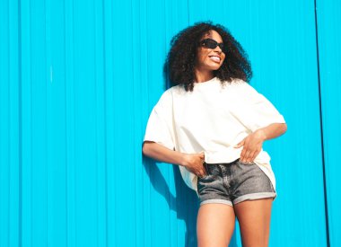 Saçları kıvırcık, güzel siyah bir kadın. Beyaz tişörtlü, hippi bir model. Sokakta mavi duvarın yanında güneş gözlükleriyle poz veren kaygısız seksi kadın. Neşeli ve mutlu