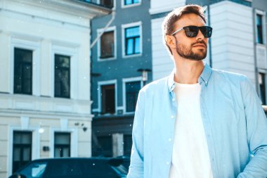 Yakışıklı, kendine güvenen, havalı, havalı, lamberseksüel model. Mavi gömlekli, modern bir adam. Moda erkeği güneş gözlükleriyle gökdelenin yanında poz veriyor. Gün batımında dışarıda.