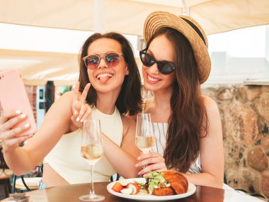 Son moda yaz kıyafetleri içinde iki güzel gülümseyen yeniyetme kadın. Sokakta Veranda Kafe 'de poz veren kaygısız kadınlar. Şapkada beyaz şarap içen pozitif modeller.