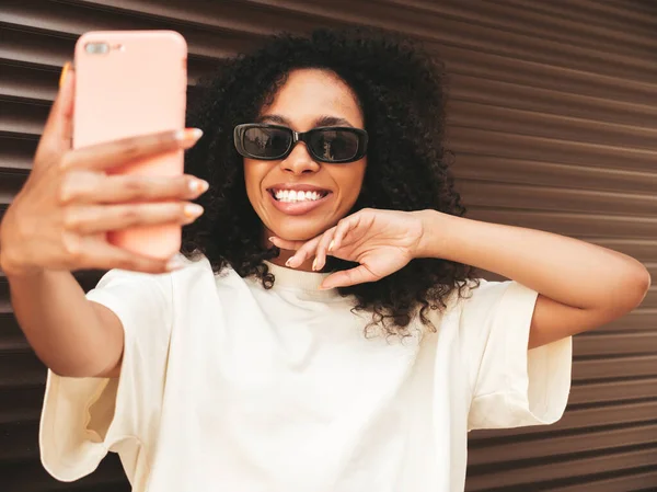 Beautiful black woman with afro curls hairstyle.Smiling hipster model in white t-shirt. Sexy carefree female posing in the street near brown wall in sunglasses. Cheerful and happy.Taking selfie