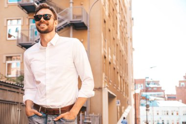 Yakışıklı, gülümseyen stil sahibi Lambersex modelinin portresi. Beyaz gömlek giymiş modern bir adam. Sokakta güneş gözlükleriyle poz veren modacı erkek. Gün batımında dışarıda. 