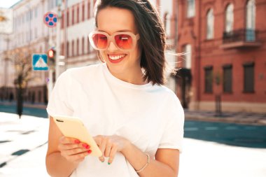 Moda kıyafetleri içinde gülümseyen genç ve güzel bir hippi kadın. Sokakta güneş gözlükleriyle poz veren kaygısız seksi kadın. Akıllı telefon uygulamaları kullanarak, ekrana bakarak pozitif model.