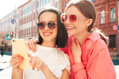Son moda beyaz tişörtlü ve kot pantolonlu iki güzel gülümseyen yeniyetme kadın. Sokakta arka planda poz veren kaygısız kadınlar. Akıllı telefon uygulamaları kullanan, ekrana bakan pozitif modeller.
