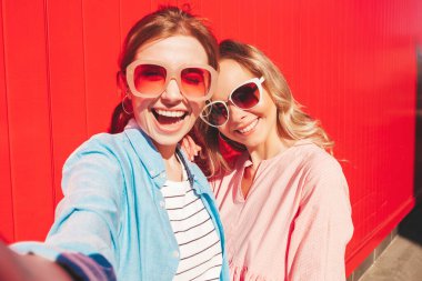 Son moda yaz kıyafetleri içinde iki güzel gülümseyen yeniyetme kadın. Kırmızı renkli duvarın yanında poz veren kaygısız kadınlar. Pozitif modeller eğleniyor, sarılıyor ve Pov 'un selfie' sini çekiyor.