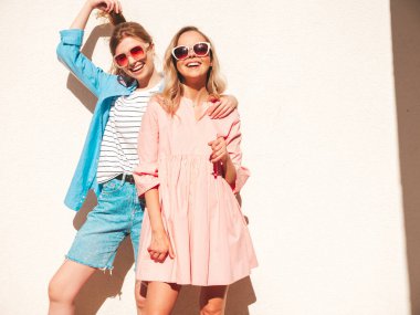 Son moda yaz kıyafetleri ve elbiseleri içinde iki güzel, gülümseyen hippi kadın. Sokakta beyaz duvarın yanında poz veren kaygısız kadınlar. Pozitif saf modeller eğleniyor, sarılıyor ve deliriyor.