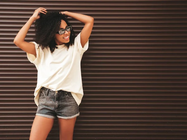 Beautiful black woman with afro curls hairstyle.Smiling hipster model in white t-shirt. Sexy carefree female posing in the street near brown wall. Cheerful and happy in sunglasses