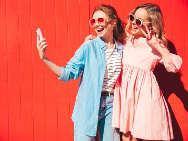 Son moda yaz kıyafetleri içinde iki güzel, gülümseyen yeniyetme kadın. Kırmızı renkli duvarın yanında sokakta poz veren kaygısız kadınlar. Pozitif modeller eğleniyor, sarılıyor ve selfie çekiyor.