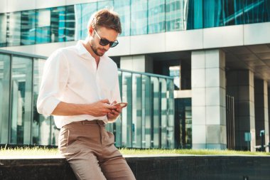 Yakışıklı, güler yüzlü, stil sahibi, lamberseksüel model. Beyaz gömlekli, modern bir adam. Moda erkek güneş gözlüklü gökdelenlerin yakınında poz veriyor. Gün batımında dışarıda. Akıllı telefon uygulamaları kullanılıyor 