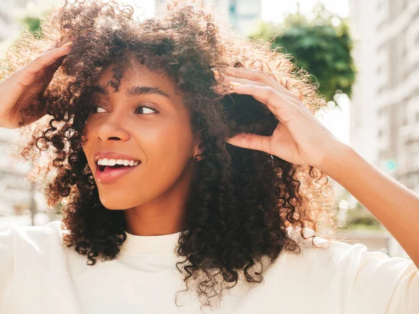 Beautiful black woman with afro curls hairstyle.Smiling hipster model in white t-shirt. Sexy carefree female posing on the street background. Cheerful and happy