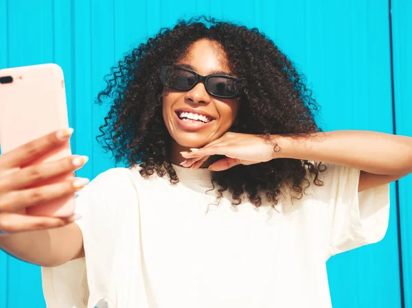 Beautiful black woman with afro curls hairstyle.Smiling hipster model in white t-shirt. Sexy carefree female posing in the street near blue wall in sunglasses. Cheerful and happy.Taking selfie photo