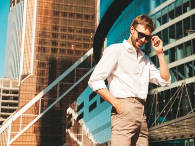 Yakışıklı, gülümseyen stil sahibi Lambersex modelinin portresi. Beyaz gömlek giymiş modern bir adam. Moda erkek, güneş gözlüklü gökdelenlerin yanında sokak arkasında poz veriyor. Gün batımında dışarıda. 