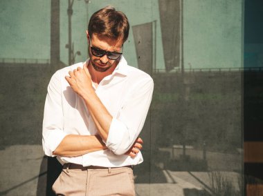 Yakışıklı, kendine güvenen, stil sahibi, Lambersex mankeni portresi. Beyaz gömlek giymiş modern bir adam. Moda erkek, güneş gözlüklü gökdelenlerin yanında sokak arkasında poz veriyor. Gün batımında dışarıda. 