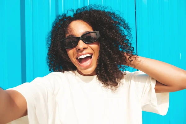 Beautiful black woman with afro curls hairstyle.Smiling hipster model in white t-shirt.Sexy carefree female posing in the street near blue wall in sunglasses.Cheerful and happy.Taking Pov selfie photo