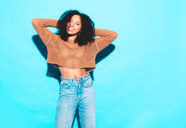 Saçları kıvırcık, güzel siyah bir kadın. Moda kıyafetleri içinde gülümseyen bir model. Stüdyoda mavi duvarın yanında poz veren kaygısız seksi kadın. Bronzlaşmış ve neşeli