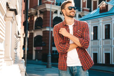 Yakışıklı, şık, havalı, havalı, lamberseksüel model. Kırmızı kareli gömlek giymiş modern bir adam. Moda erkeği güneş gözlükleriyle gökdelenin yanında poz veriyor. Gün batımında dışarıda. 