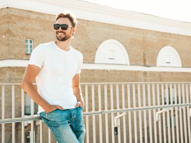Yakışıklı, gülümseyen hipster Lambersex modelinin portresi. Beyaz tişört ve kot giyen şık bir adam. Moda erkek, güneş gözlüklü, plajda çitlerin yanında poz veriyor.