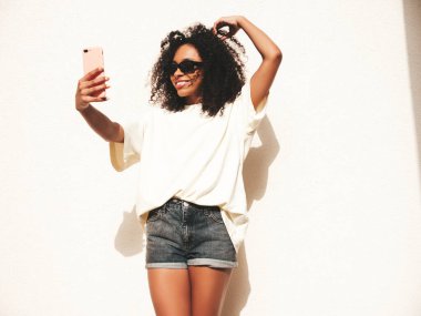 Saçları kıvırcık, güzel siyah bir kadın. Beyaz tişörtlü, hippi bir model. Sokakta güneş gözlüklü beyaz duvarın yanında poz veren kaygısız seksi kadın. Neşeli ve mutlu. Selfie çekiyoruz.