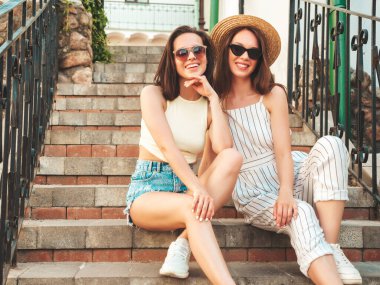Son moda yaz kıyafetleri içinde iki güzel gülümseyen yeniyetme kadın. Sokakta şapkalı kayla poz veren kaygısız kadınlar. Pozitif saf modeller gün batımında eğleniyorlar, merdivenlerde oturuyorlar.
