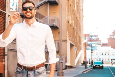 Yakışıklı, kendine güvenen, stil sahibi, Lambersex mankeni portresi. Beyaz gömlek giymiş modern bir adam. Sokakta güneş gözlükleriyle poz veren modacı erkek. Gün batımında dışarıda. 