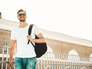 Yakışıklı, güler yüzlü, Lambersex mankeni. Beyaz tişört ve kot giyen şık bir adam. Moda erkek plajda çitlerin yanında güneş gözlüklü okul çantasıyla poz veriyor.