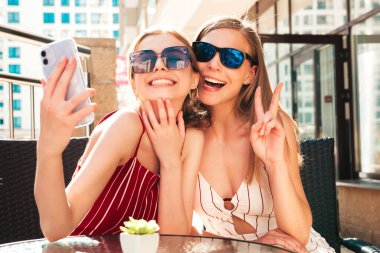 Son moda yaz kıyafetleri içinde iki güzel, gülümseyen yeniyetme kadın. Sokakta Veranda Kafe 'de poz veren kaygısız kadınlar. Pozitif modeller eğleniyor ve akıllı telefonlarda selfie çekiyor.
