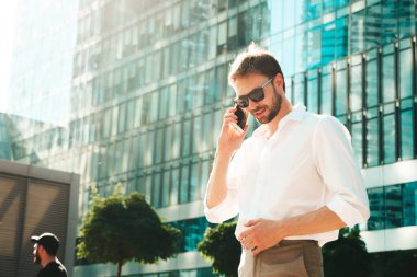 Yakışıklı, güler yüzlü, stil sahibi, lamberseksüel model. Beyaz gömlekli, modern bir adam. Sokakta güneş gözlükleriyle poz veren modacı erkek. Gün batımında dışarıda. Akıllı telefon kullanarak. Konuşarak.